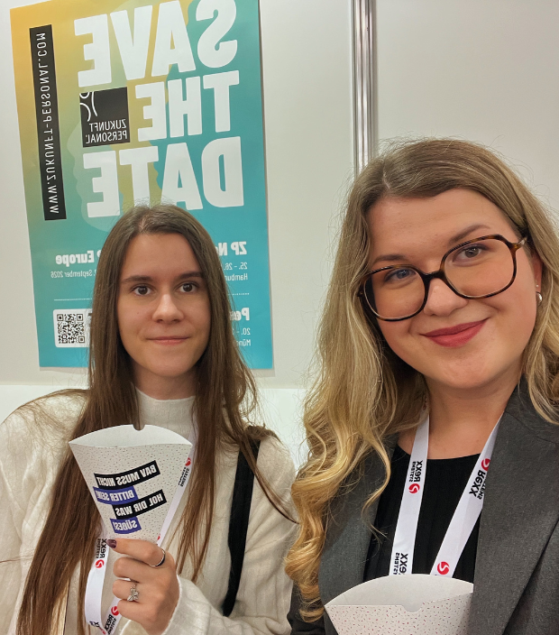 Lorena Heer und Eliza Radzele auf der Zukunft Personal 2025 vor einem Save the Date Plakat mit Messeschildern und Snacks in der Hand