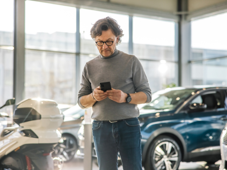 Kunde nutzt Smartphone im Autohaus zwischen Neuwagen und Motorrädern zur digitalen Information und Kundenansprache